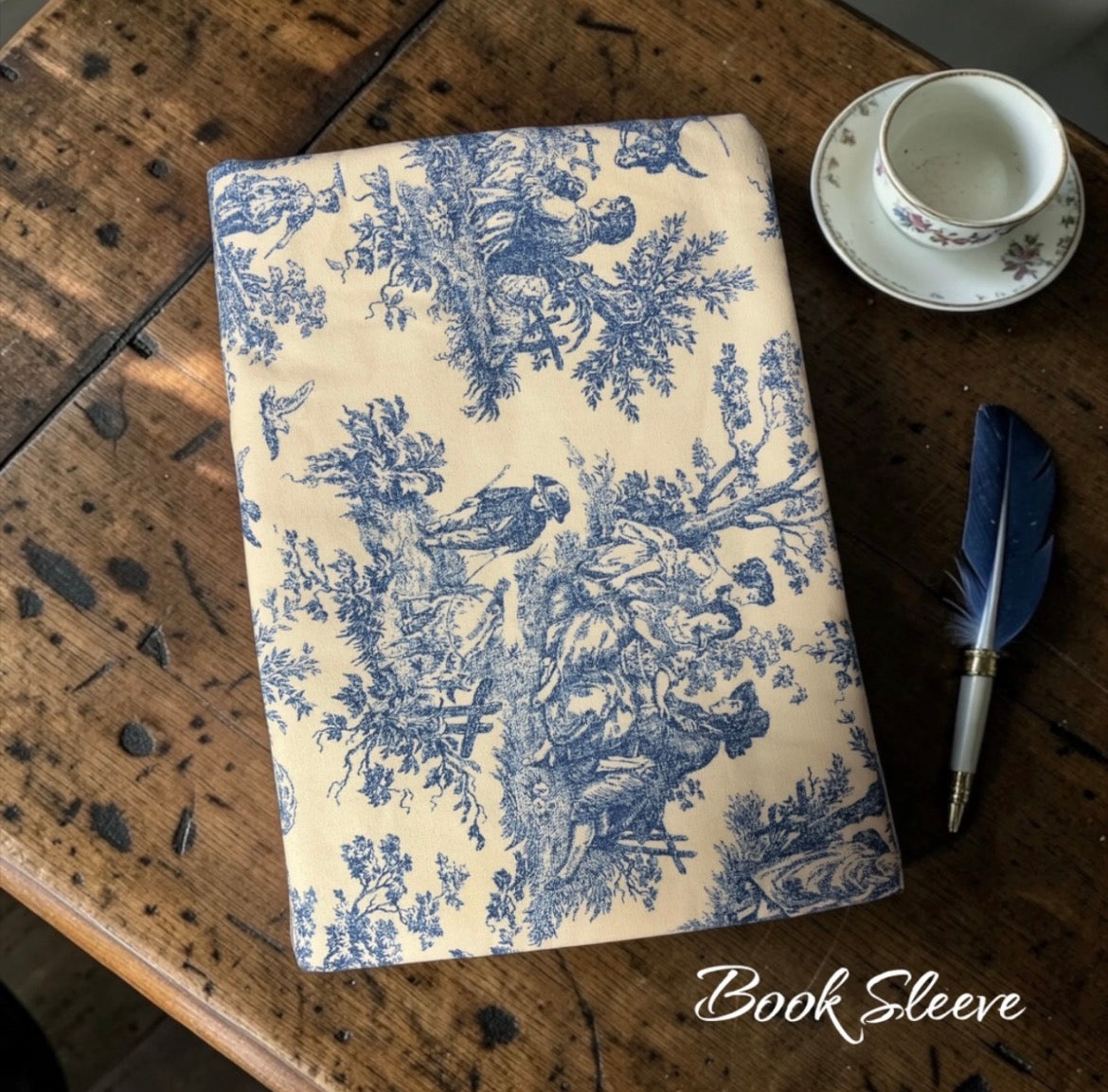 Book sleeve with blue floral pattern on a wooden surface with a cup and feather.